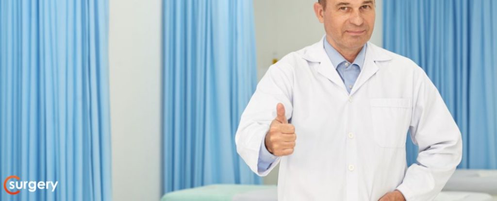 באיזה גיל מקבלים קילה? - רופא עם קרחת וחלוק לבן עומד מביט למצלמה ומסמן עם האגודל שלו. בצד שמאל למטה לוגו אתר surgery