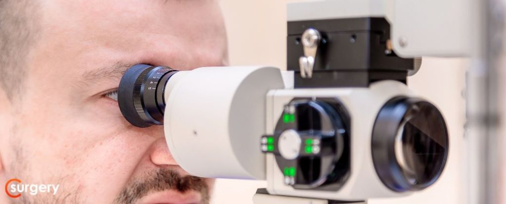 חודש אחרי ניתוח קטרקט: איש מקצוע רפואי עם חלוק כחול מתבונן במכשיר לבדיקת עיניים. בצד שמאל למטה לוגו אתר surgery