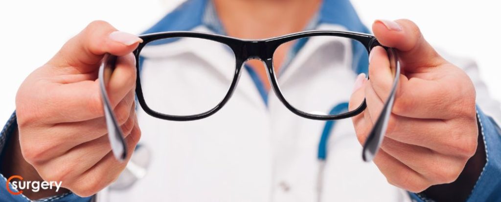 אישה עם חלוק לבן וצווארון כחול מציגה משקפיים אחרי ניתוח קטרקט. בצד שמאל למטה לוגו אתר surgery