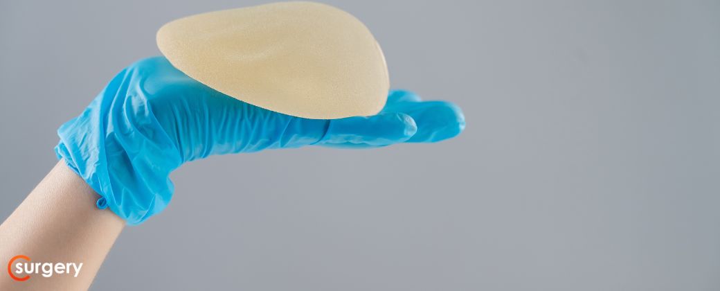 ניתוח להחלפת שתלים - באיזה מקרים: יד עם כפפה כחולה מציגה שתל סיליקון. בצד שמאל למטה לוגו אתר surgery